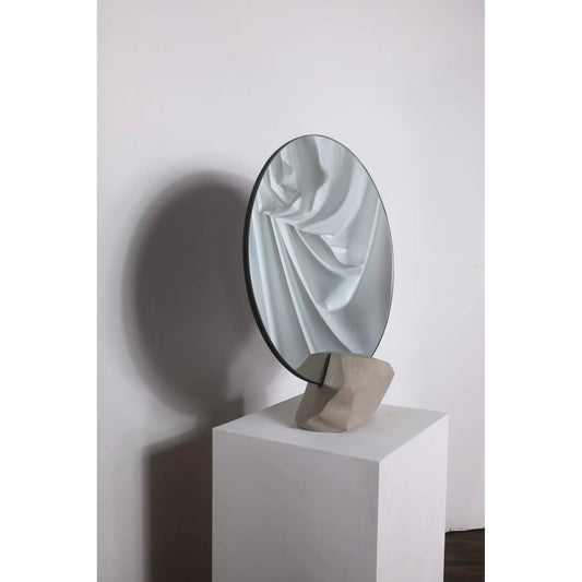 Round mirror on a pedestal with draped fabric reflection against a white wall